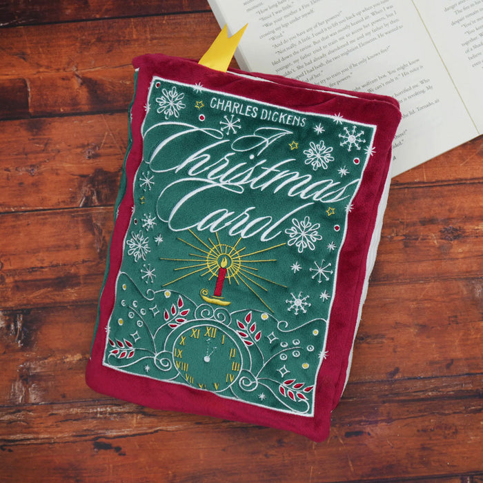 An embroidered book cover for A Christmas Carol by Charles Dickens, featuring a candle and clock design, resting on a wooden surface next to an open book.