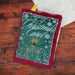 An embroidered book cover for A Christmas Carol by Charles Dickens, featuring a candle and clock design, resting on a wooden surface next to an open book.