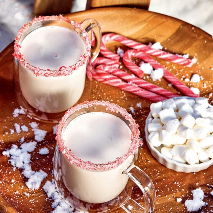 Two Christmas Cocktails in glass mugs with creamy beige drinks and crushed candy cane rims, served on a wooden tray with candy canes and a dish of marshmallows.