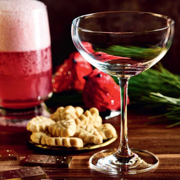 An empty stemmed cocktail glass sits on a wooden table beside a tall Christmas Cocktails drink, a plate of leaf-shaped cookies, two red birds, and dominoes.
