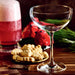 An empty stemmed cocktail glass sits on a wooden table beside a tall Christmas Cocktails drink, a plate of leaf-shaped cookies, two red birds, and dominoes.