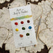Paint your own Holiday Decorations watercolor palette card for DIY crafts, featuring eight labeled paint dots, a small brush, and several gold-outlined paper ornaments.