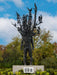 A sculpture of a human figure with branches and birds extending from its upper body stands on a pedestal outdoors at Frederik Meijer Gardens, enveloped by lush greenery. A barcode is visible on the base. The partly cloudy sky enhances the tranquil atmosphere—an ideal scene highlighted in the Guide to Collections & Gardens for anyone exploring the sculpture park.