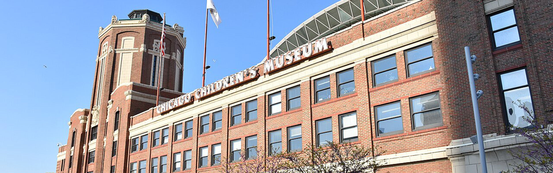 A brick building with large windows and a tower, featuring the sign Chicago Childrens Museum on the facade, under a clear blue sky with two flagpoles in front.