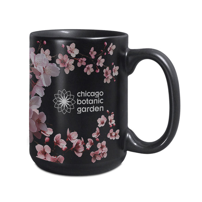 The Chicago Botanic Garden Cherry Blossom Mug is a black ceramic mug adorned with delicate pink cherry blossoms and “Chicago Botanic Garden” printed in white, alongside a floral logo. Elegant flowers are gracefully scattered around the mug, primarily on the left side and partially wrapping around for a sophisticated look.