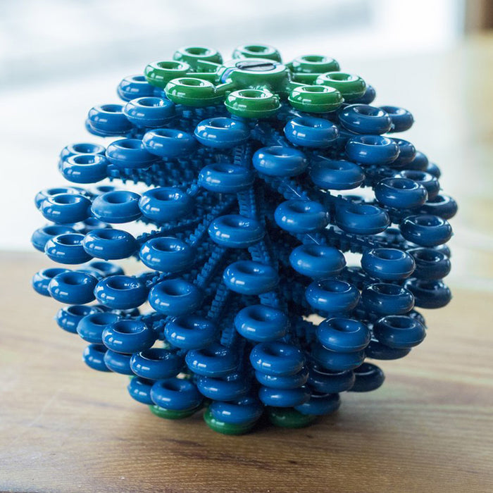 A close-up of the Cora Laundry Ball, made of interlocking blue and green plastic rings layered together, sits on a wooden surface with a blurred background. This product captures microfibers during laundry cycles.