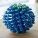 A close-up of the Cora Laundry Ball, made of interlocking blue and green plastic rings layered together, sits on a wooden surface with a blurred background. This product captures microfibers during laundry cycles.