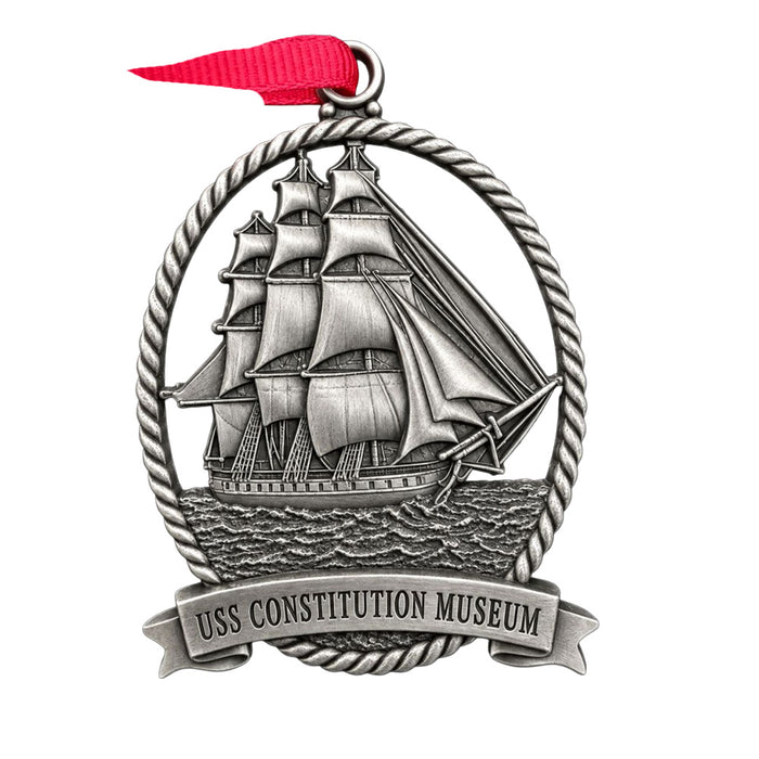 USS Constitution Pewter Ship Ornament depicting a sailing vessel inside a rope-style oval frame, topped with a red ribbon and a banner at the bottom reading USS CONSTITUTION MUSEUM.